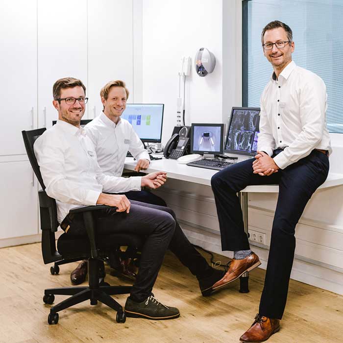 Dr. med. Nicolai Krasny, Privatdozent Dr. med. Tilman Hickethier und Dr. med. Andrej Krasny sitzen vor Bildschirmen mit radiologischen Aufnahmen und lachen freundlich in die Kamera.
