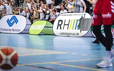 Eine Bandenwerbung der Radiologie Heinrichsallee bei einem Basketballspiel. Im Hintergrund: jubelnde Fans